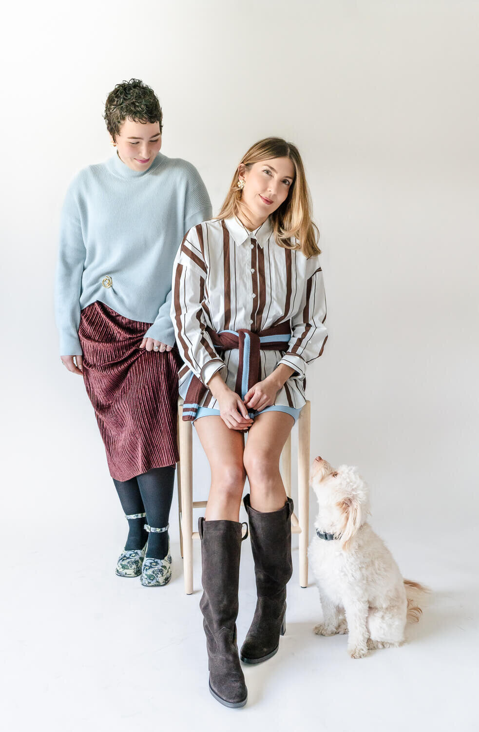 Two women and small white dog posing together in styled outfits during studio brand photoshoot.