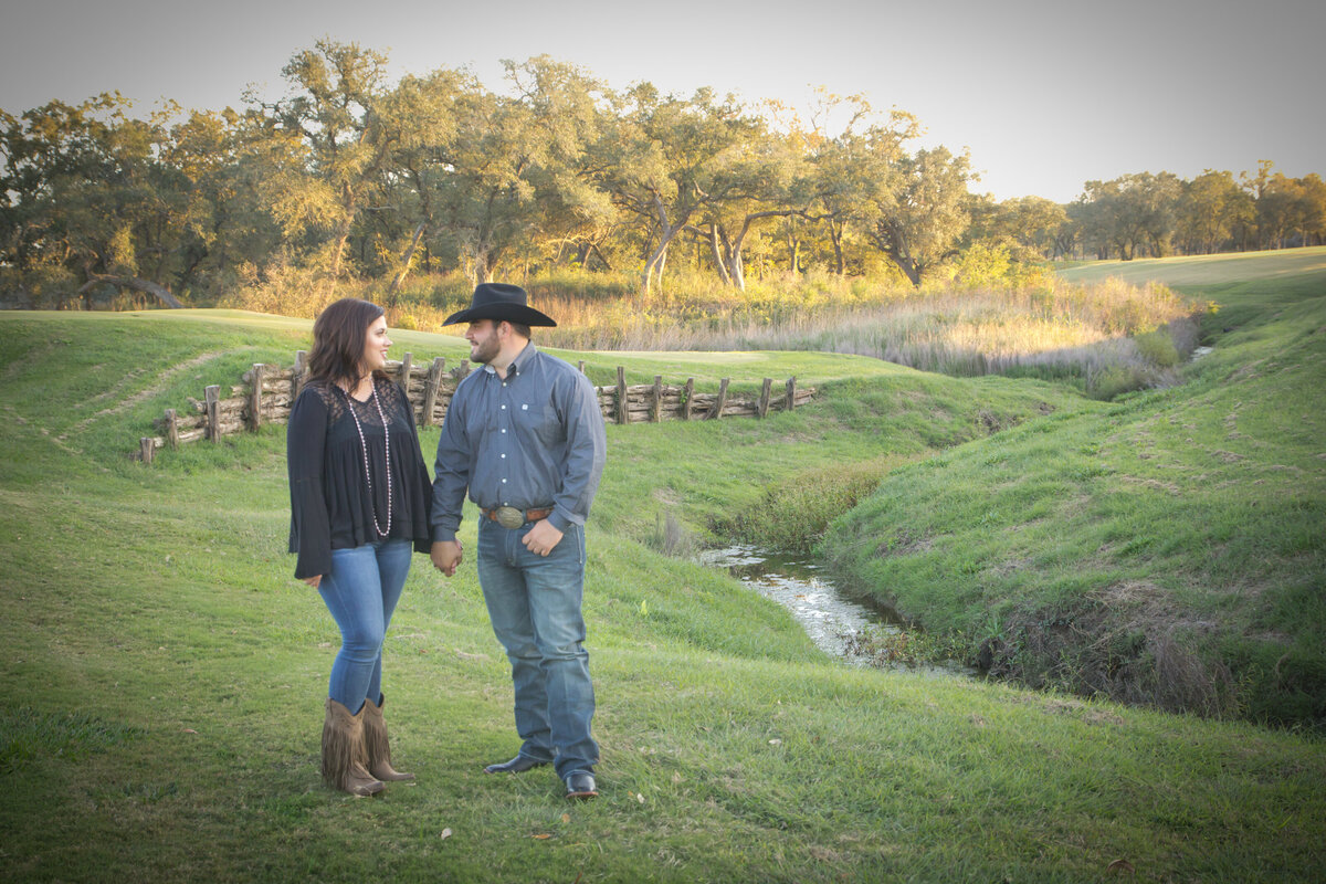 man in cowboy hat holds hands with woman by stream and rolling hills at sunset by Texas wedding photographer Firefly Photography