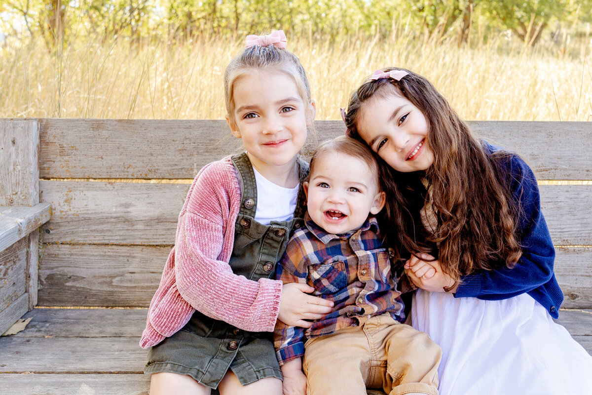 Three young siblings sit closely on a weathered wooden bench, embracing each other with wide smiles. The golden autumn scenery adds a cozy and nostalgic feel to their sweet connection.