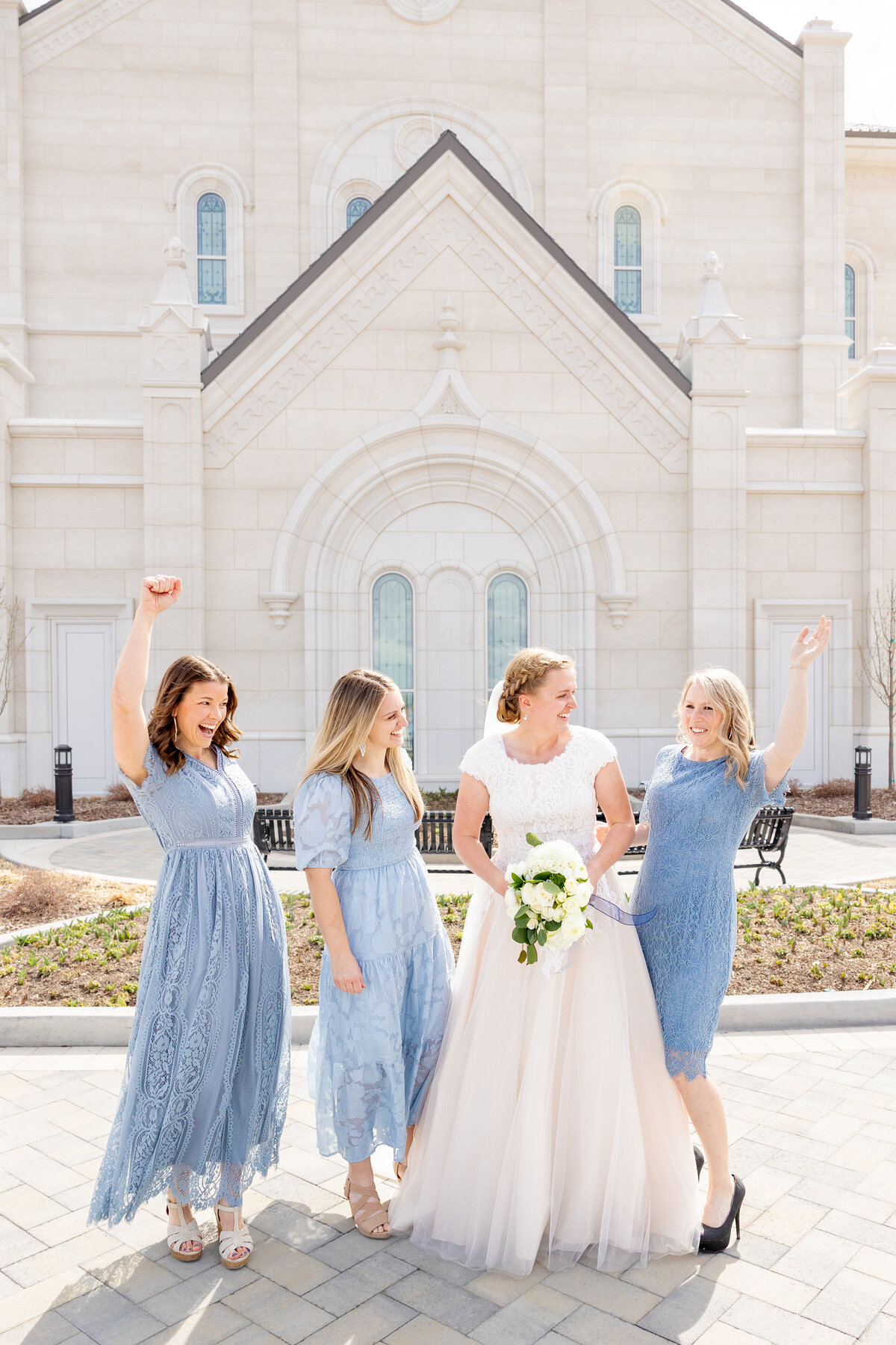 The bride and her bridesmaids cheer with raised hands in front of the LDS temple, sharing laughter and excitement.