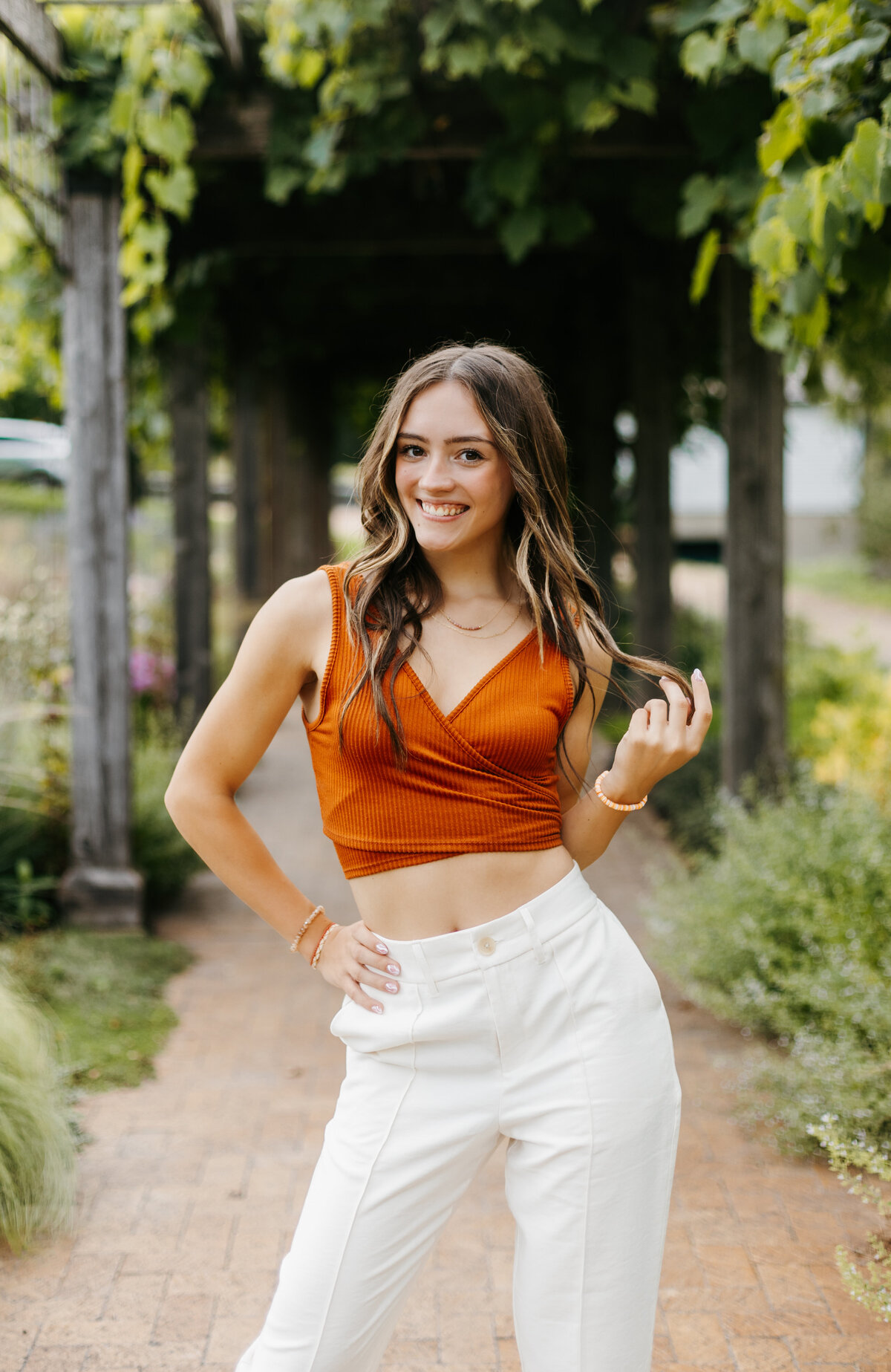 Senior girl in an orange top and white pants posing with her hand on her hip in front of a wooden arch with greenery
