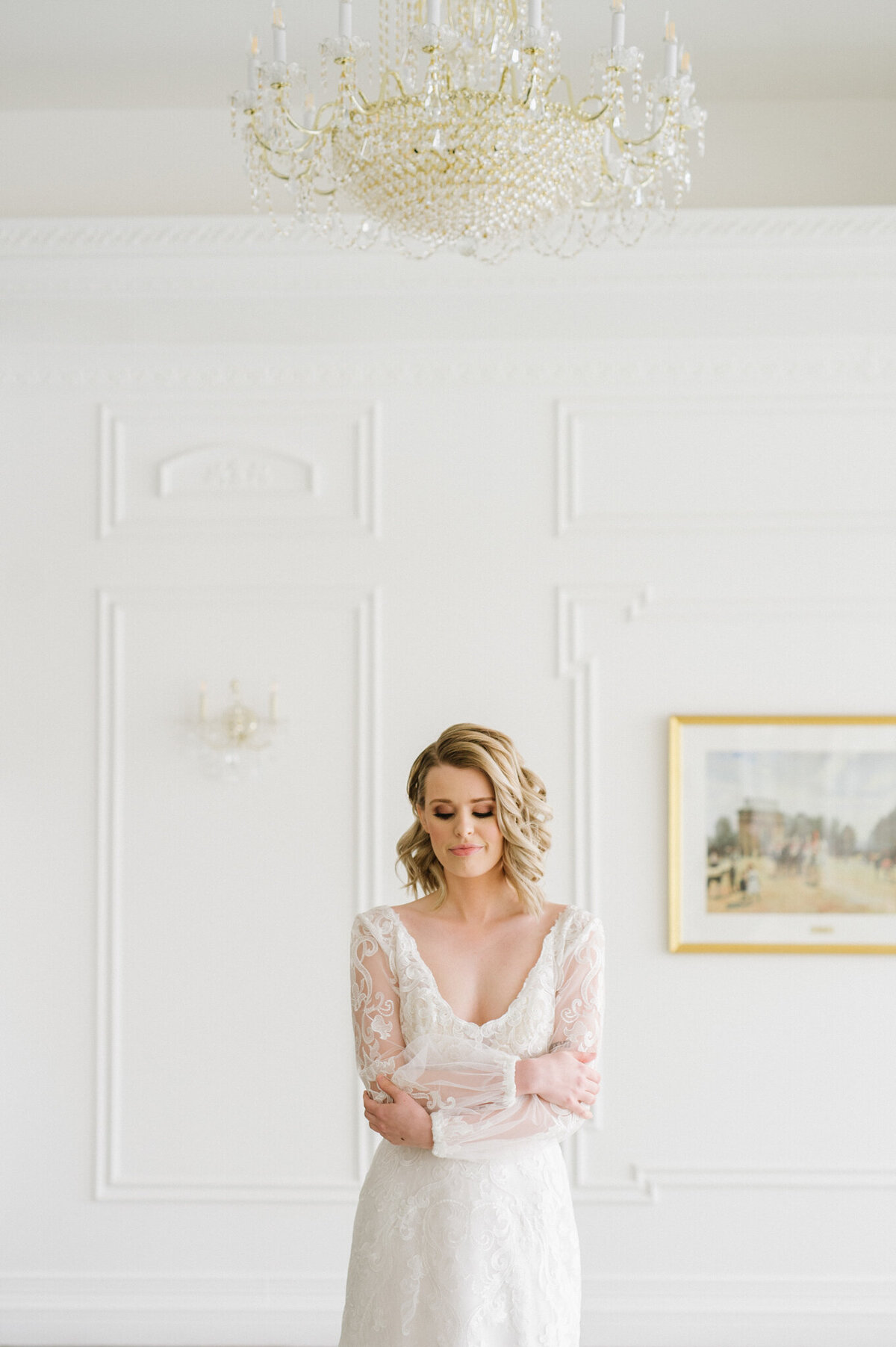 Editorial style bridal portrait, bride wearing a stunning long-sleeved gown with embroidered lace details, short wedding hair inspiration, in a white room with a gorgeous chandelier, captured by Christy D. Swanberg Photography, editorial elopement and wedding photographer in Calgary, Alberta, featured on the Bronte Bride Vendor Guide.