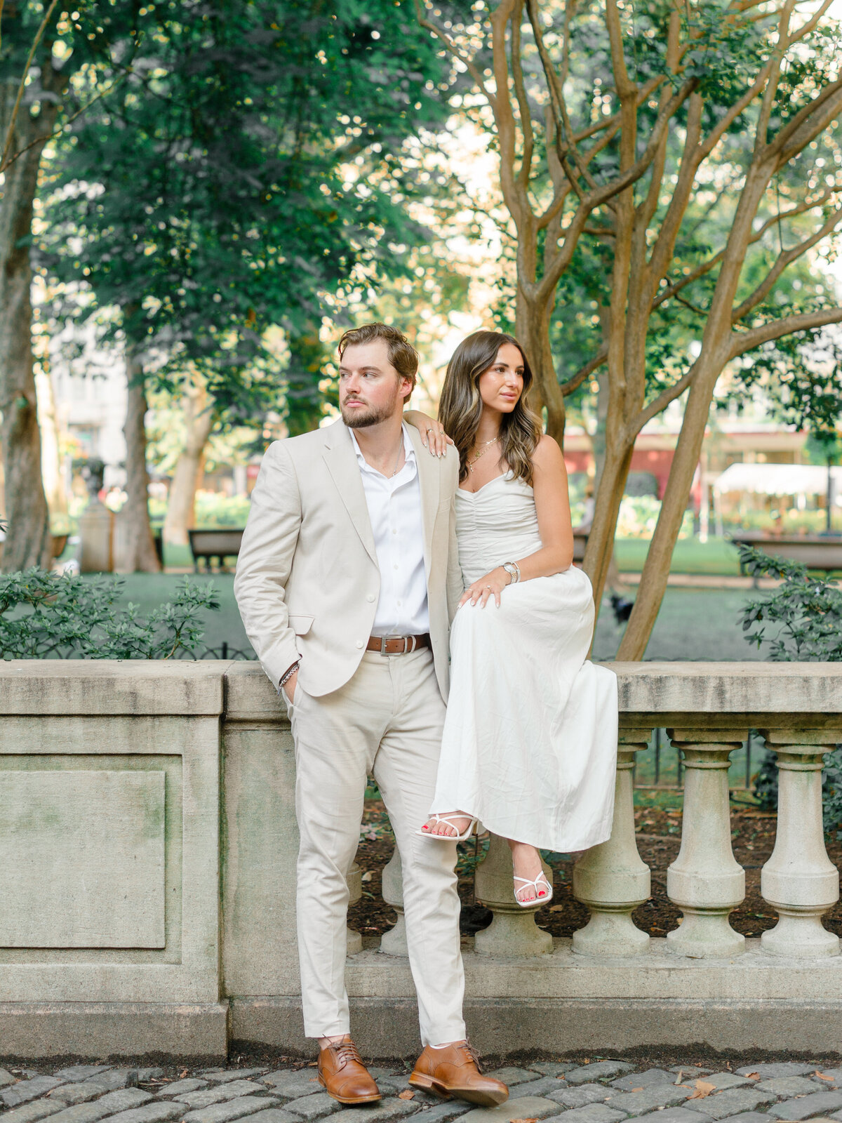A fashionable couple poses along a stone balustrade in Rittenhouse Square, surrounded by tall trees and classic garden charm. The woman sits gracefully in a white dress while resting her hand on her partner’s shoulder, both gazing thoughtfully into the distance. Captured by Elizabeth Kane Photography, a Luxury Philadelphia Wedding Photographer.