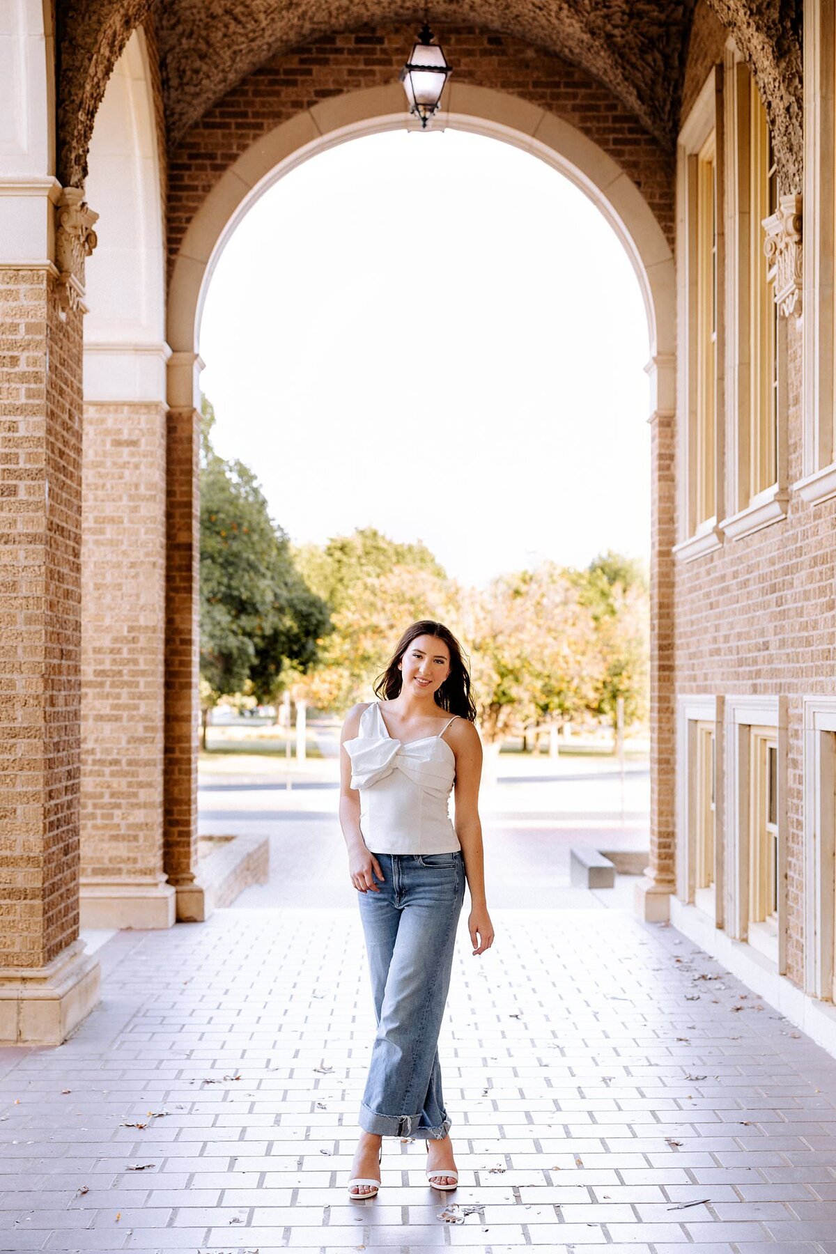 Senior in denim and white walking under an arched building in Lubbock Texas.