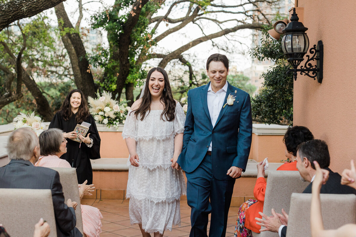 A bride and groom joyously walking down the aisle after their wedding ceremony at the Rosewood Mansion on Turtle Creek in Dallas, Texas. The bride is on the left and is wearing a short, intricate, white dress. The groom is on the right and is wearing a blue suit with a boutonniere. Their officiant is standing and smiling behind them off to the side while many wedding guests applaud and cheer from their seats on either side of the aisle.