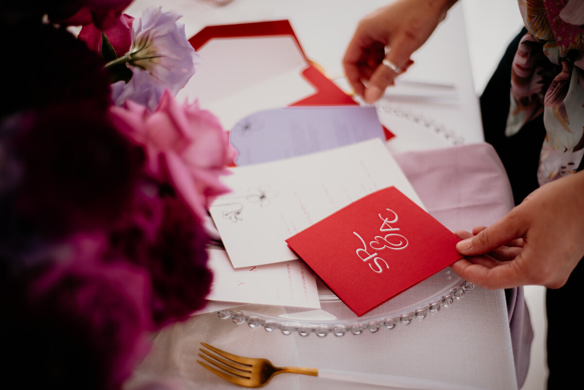 Two hands holding a bright red and purple wedding invitation with contemporary monogram