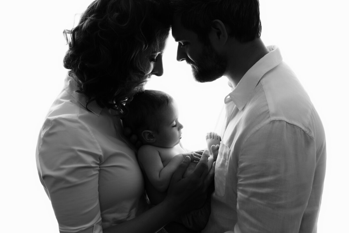 Mother and father dressed in white tenderly looking at their newborn that they are holding between them.