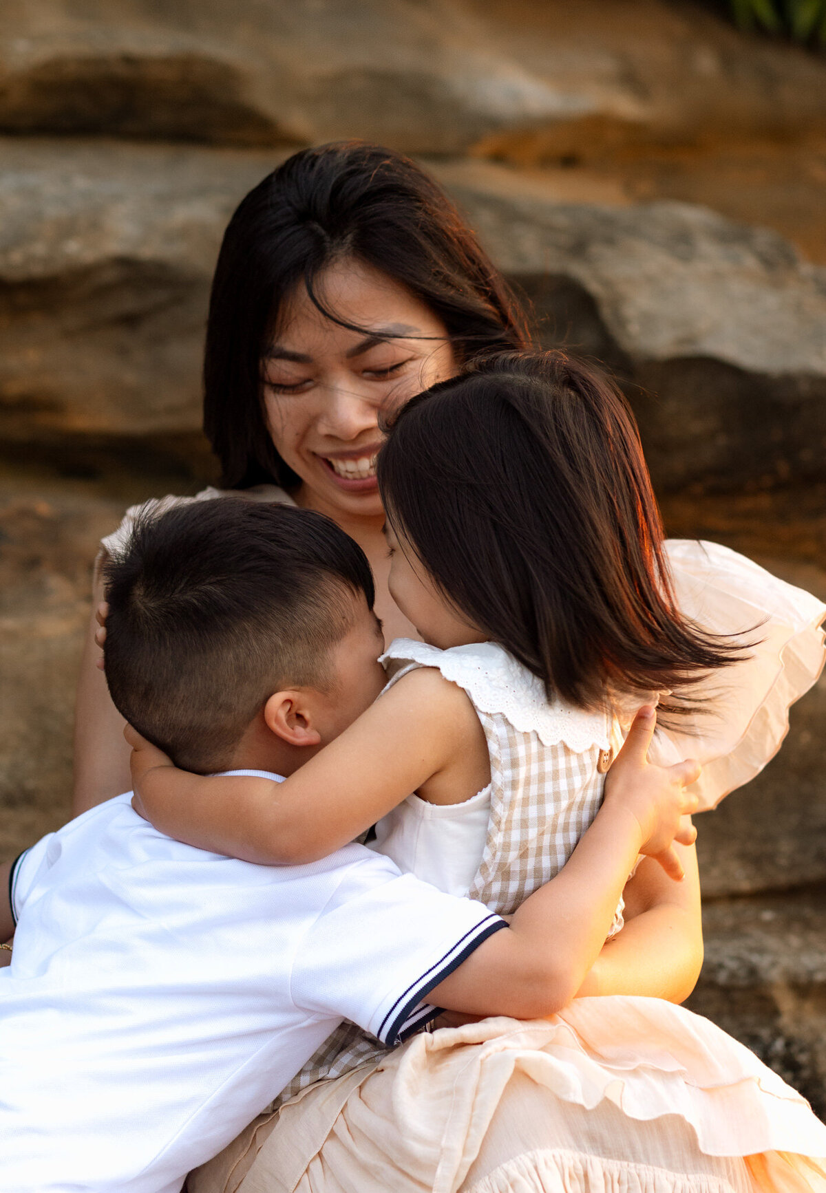 Mother embracing her two young children near sandstone rocks at sunset, all smiling and sharing a joyful hug during a candid family photography session.