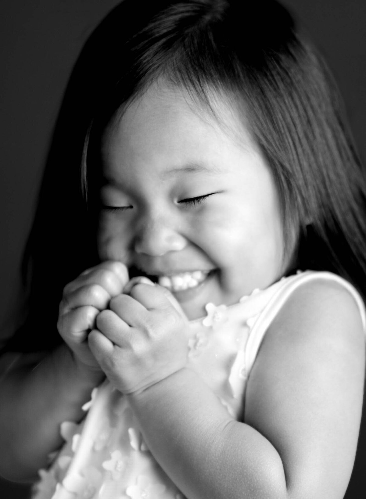 A black and white photo of a young girl smiling with joy. Her eyes are closed and hands clasped near her face, wearing a textured top. The mood is playful and happy.