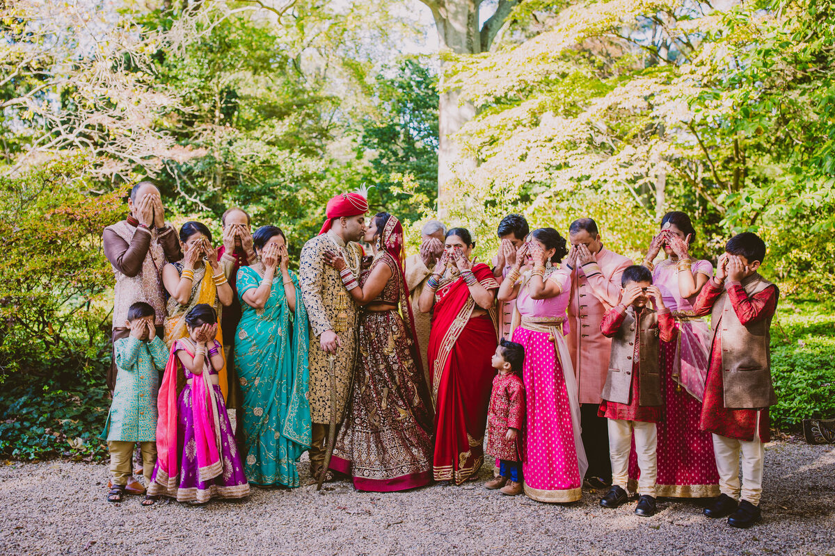 Family covering their eyes during the portraits of the indian bride and groom kissing at the planting fields arboretum