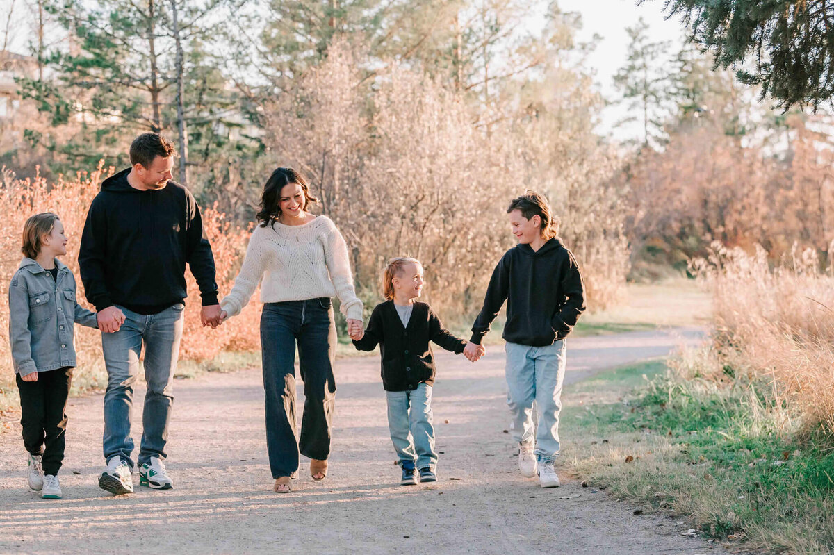 The family of 5 is holding hands  and walking down a path along the river is Saskatoon. 