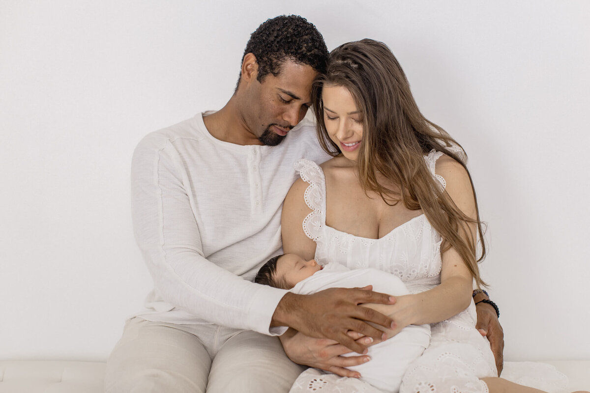 A couple sits closely together on a white couch, both wearing light-colored clothing. They are gently holding a sleeping baby wrapped in a white blanket, with warm expressions, creating a serene and loving family moment.