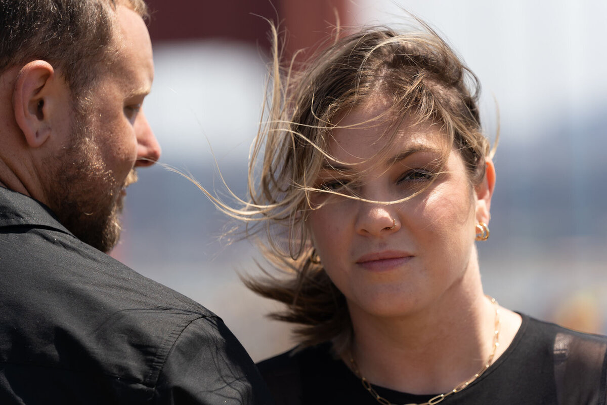 Close up photo of a couple embracing with significant wind and the golden gate bridge in the background.