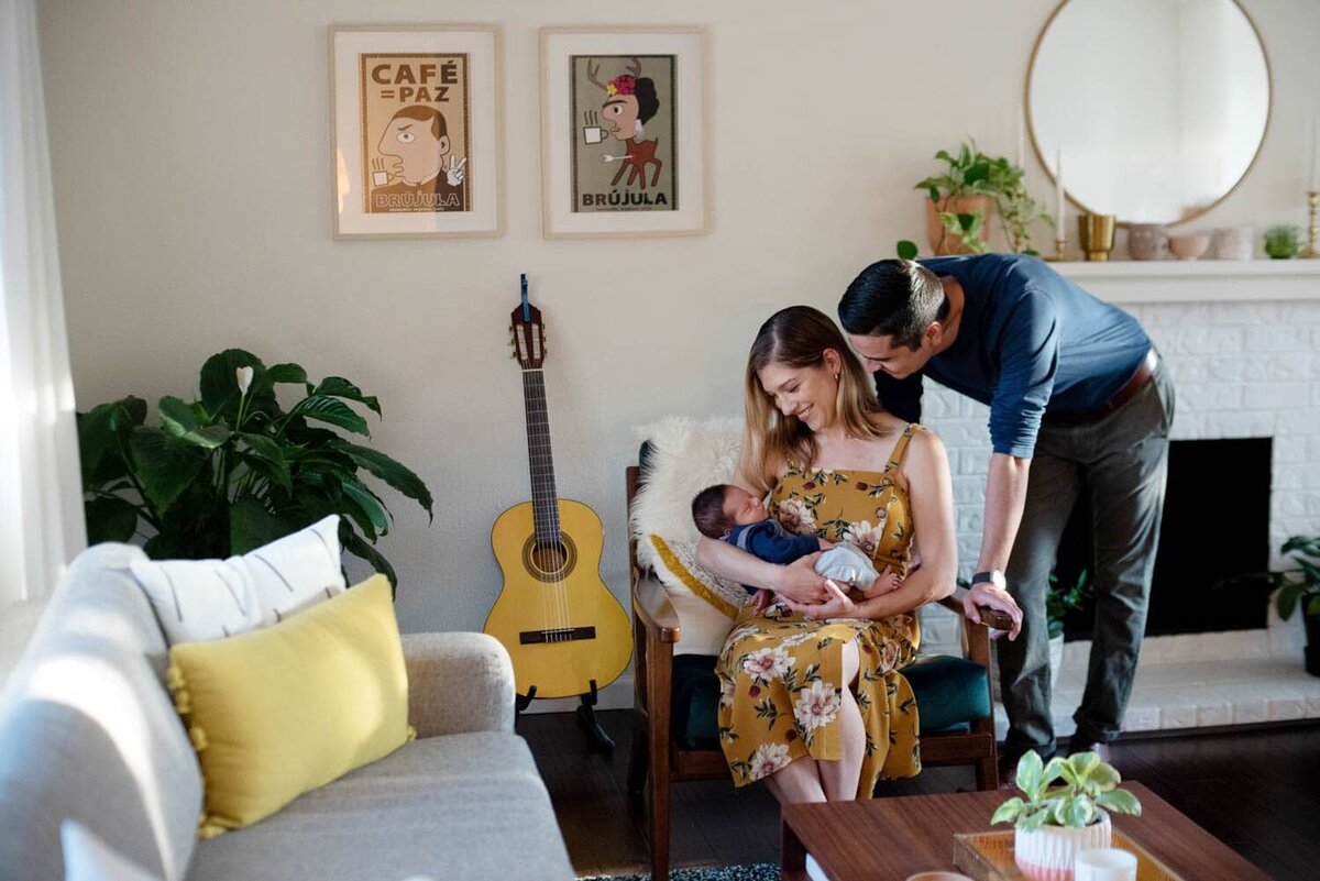 New mother in a yellow floral dress sits in colorful house with the baby while dad leans over to see baby,