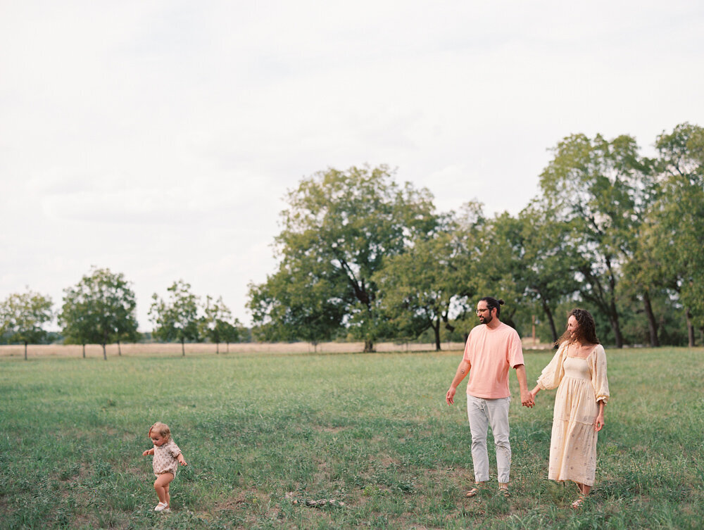 Mom and dad holding hands in a grassy field watching their baby walking through the grass