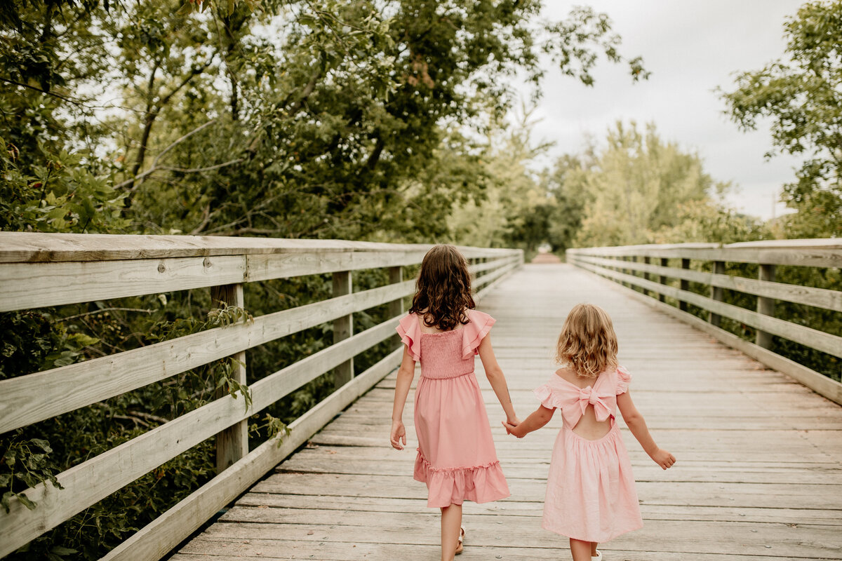 Sisters wearing pink dresses walk along a wooden bridge holding hands during summer family photos.