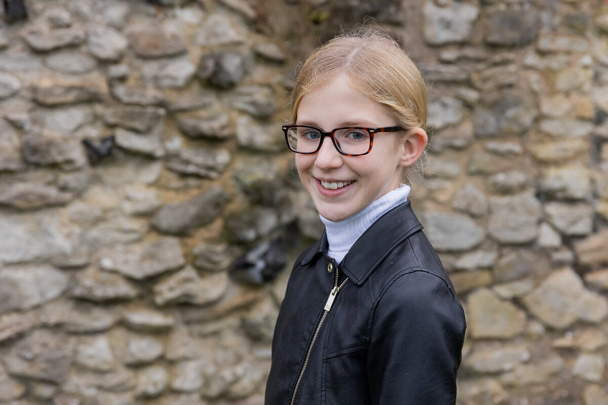 A young girl with glasses smiling at the camera, standing in front of a stone wall. she wears a black jacket and a white turtleneck.