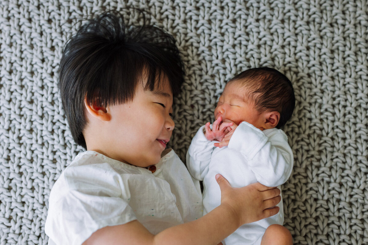 brother laying on textured blanket next to newborn sister and looking at her