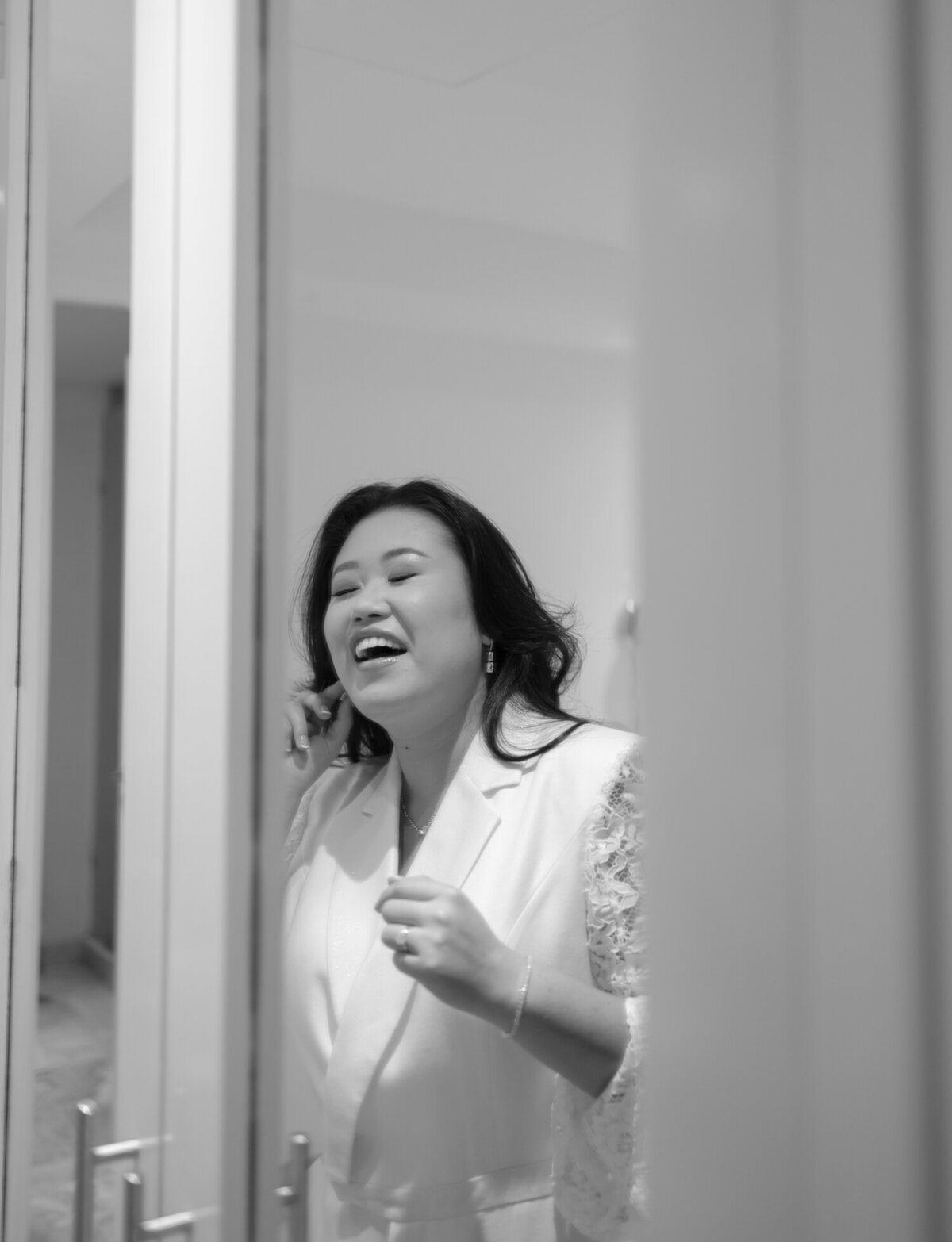 asian bride in chic pantsuit laughs in mirror, putting earrings on
