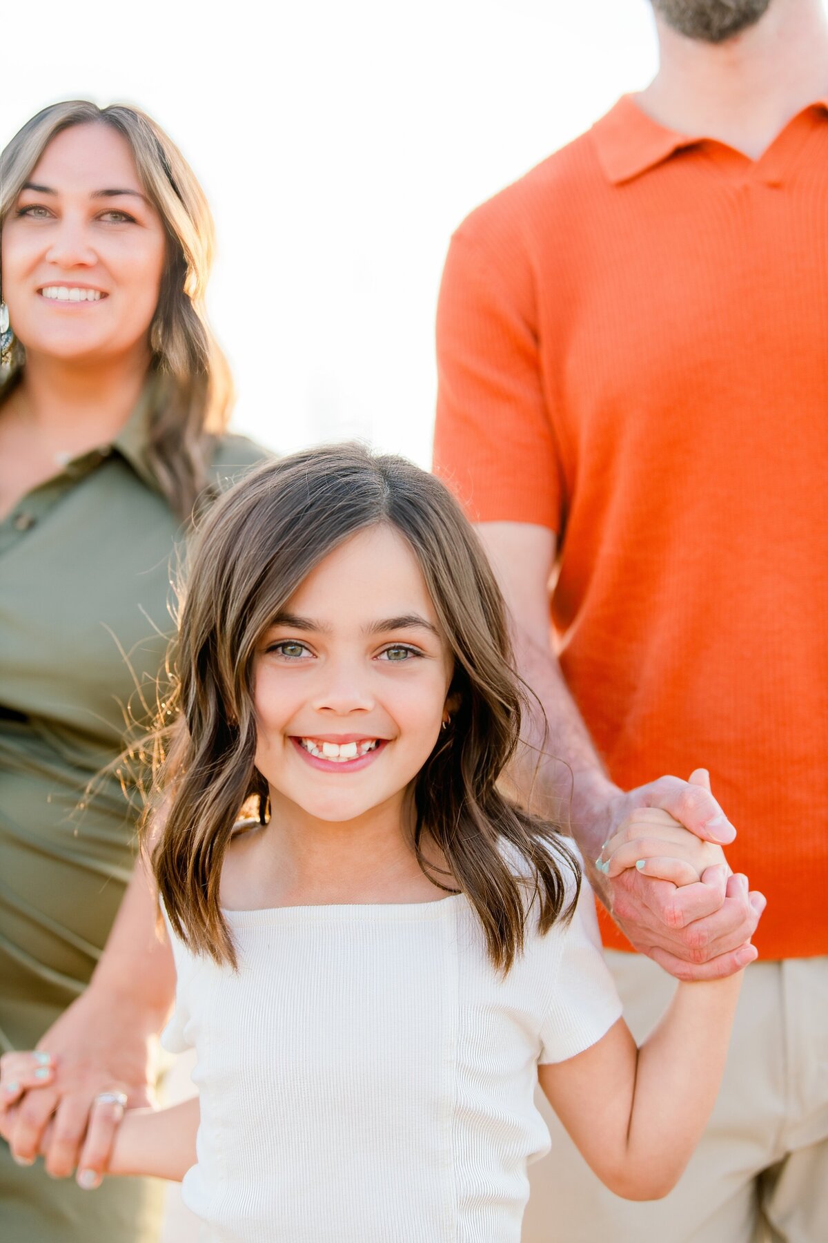 little girl smiling at the camera holding her parents hands