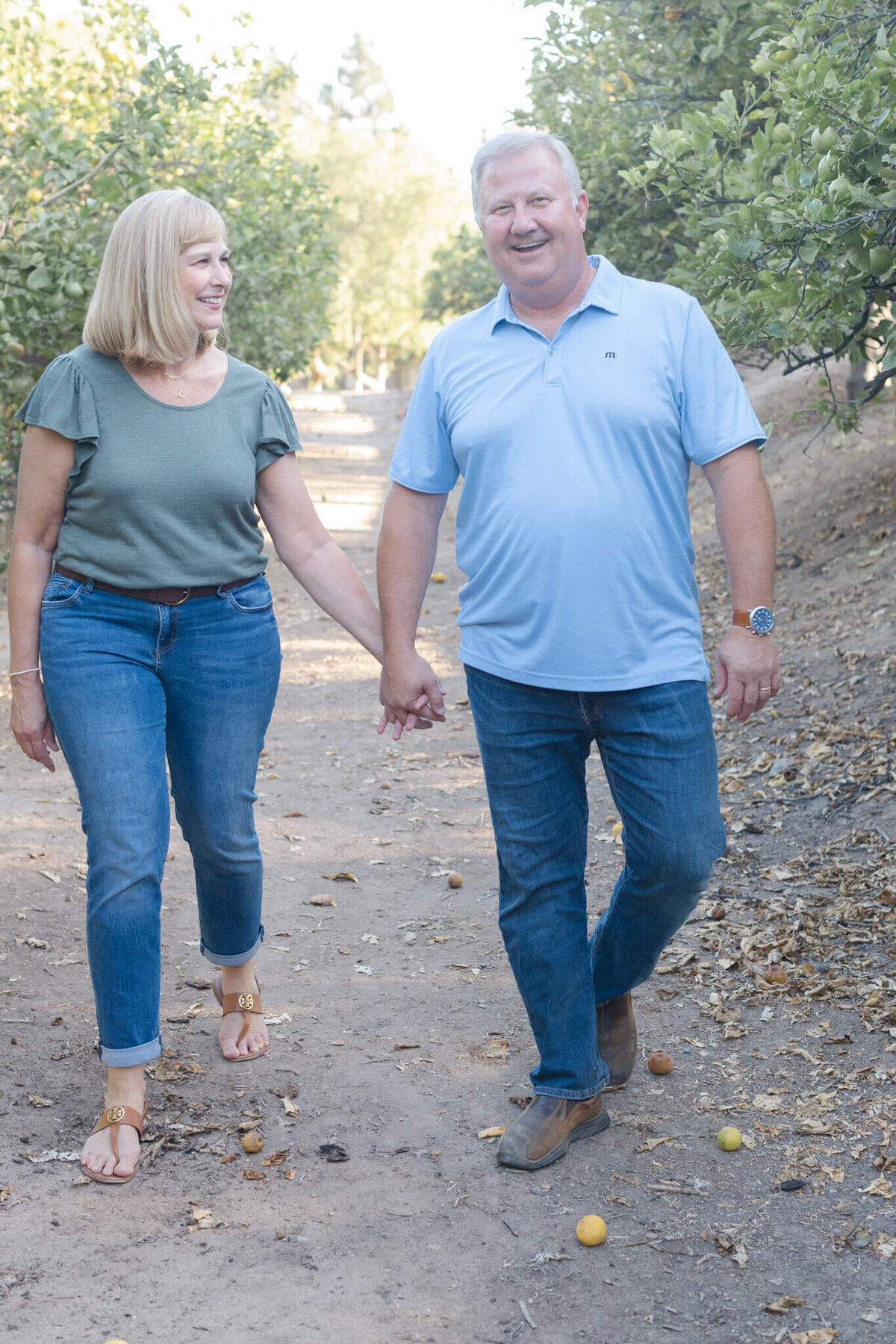 A husband and wife holding hands, walking towards the camera as husband is smiling at the camera and his wife is smiling at him.