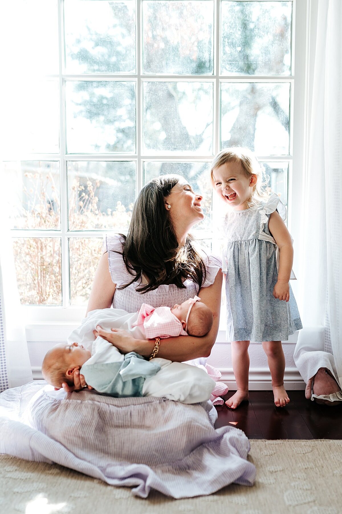 mother sitting on floor holding newborn twins laughing at toddler daughter