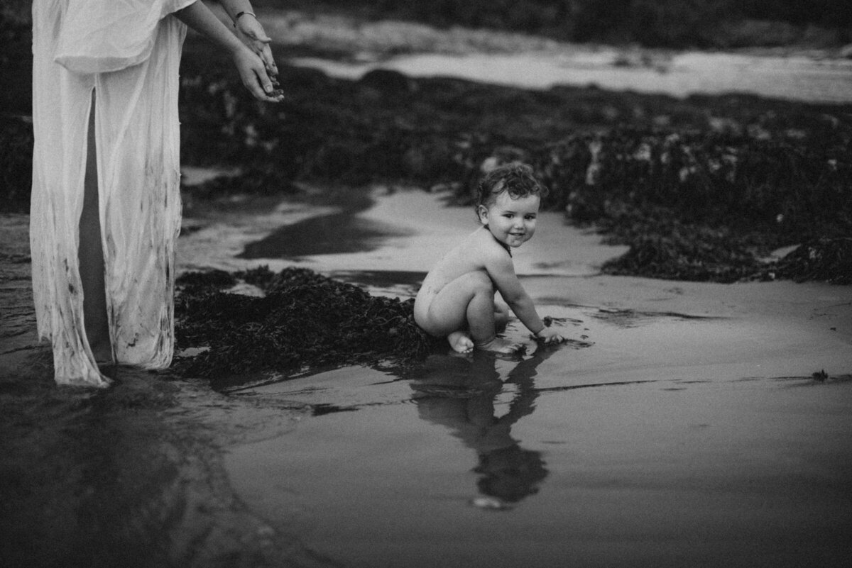 Black and White image of baby playing in the sand while mother looks on in Maine