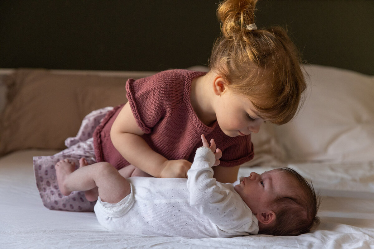 Livsstilsfoto av to små barn på en seng; storesøster på ett år lener seg over babyen som ligger på rygg og ser på storesøster.