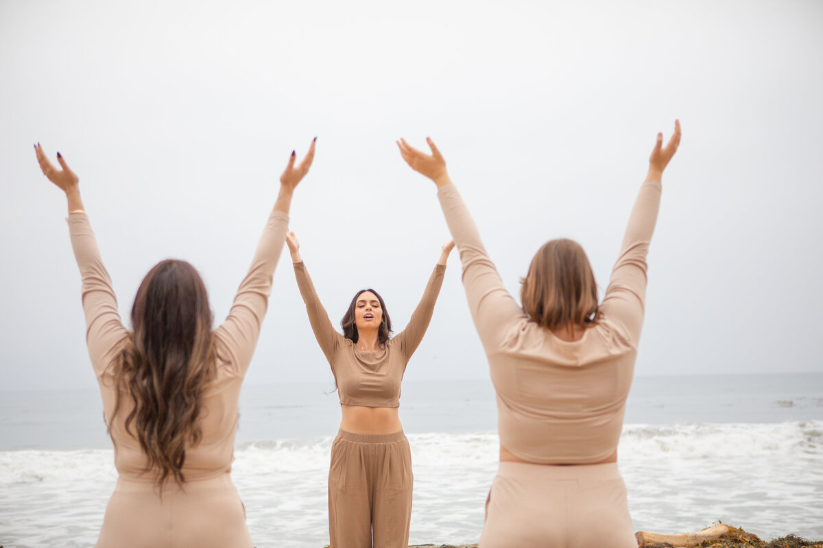 Three women wearing matching neutral-toned outfits raise their arms in a synchronized pose during a mindful movement session on the El Matador beach, with waves crashing behind them.