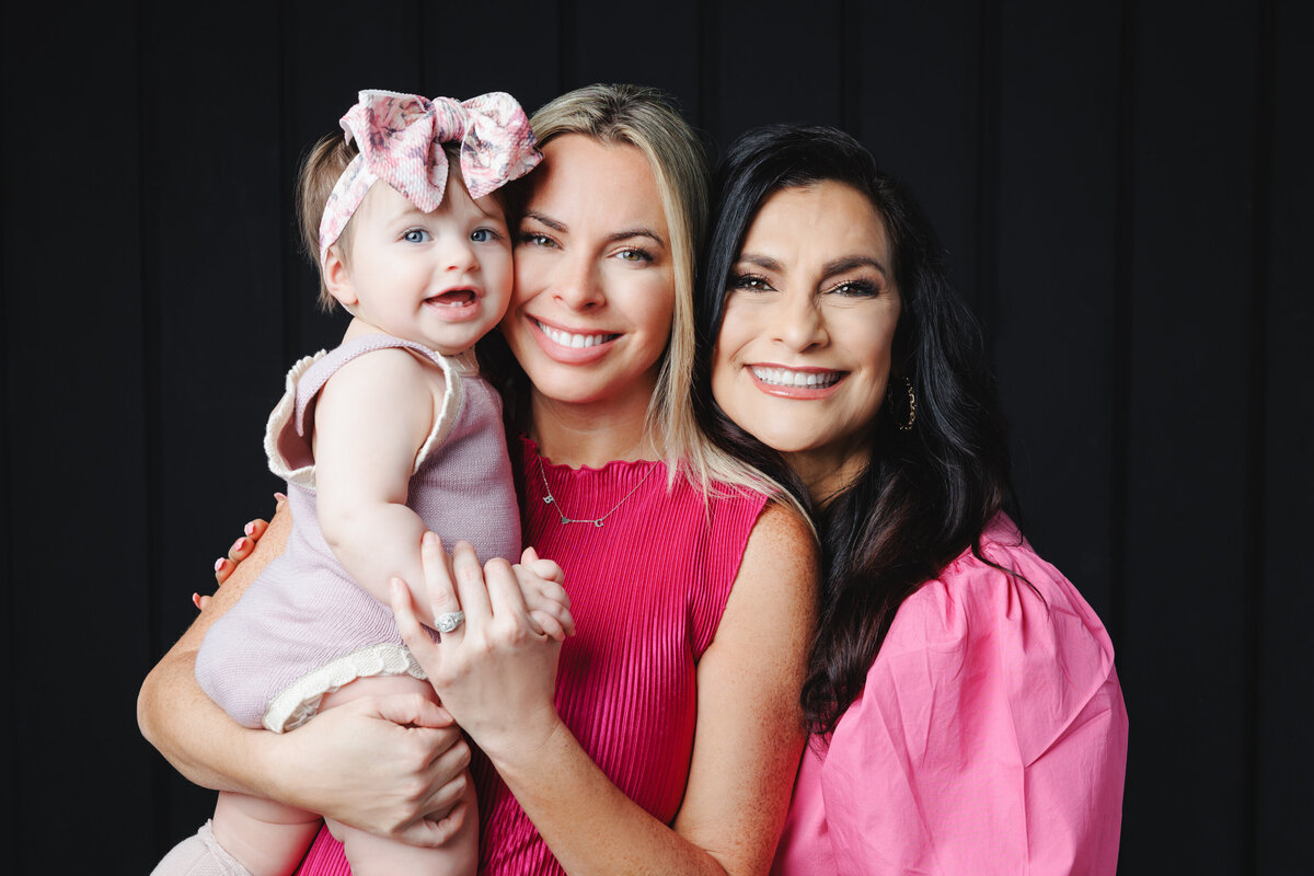 Portrait of two women and a baby all wearing pink posed in front of a black backdrop