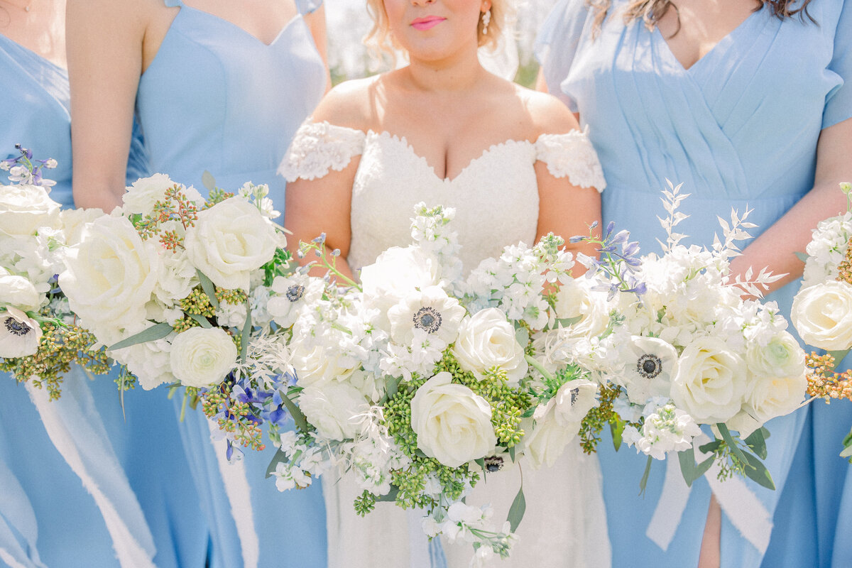 Bride in her wedding dress with bridesmaids in their blue dresses holding their bouquets of white florals and greenery