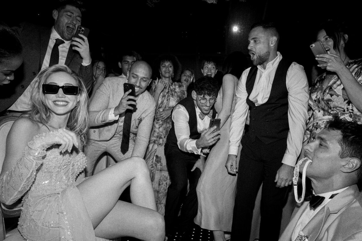 A destination wedding photo of a groom taking the garter out of a brides leg iat the party with guests surrounding them