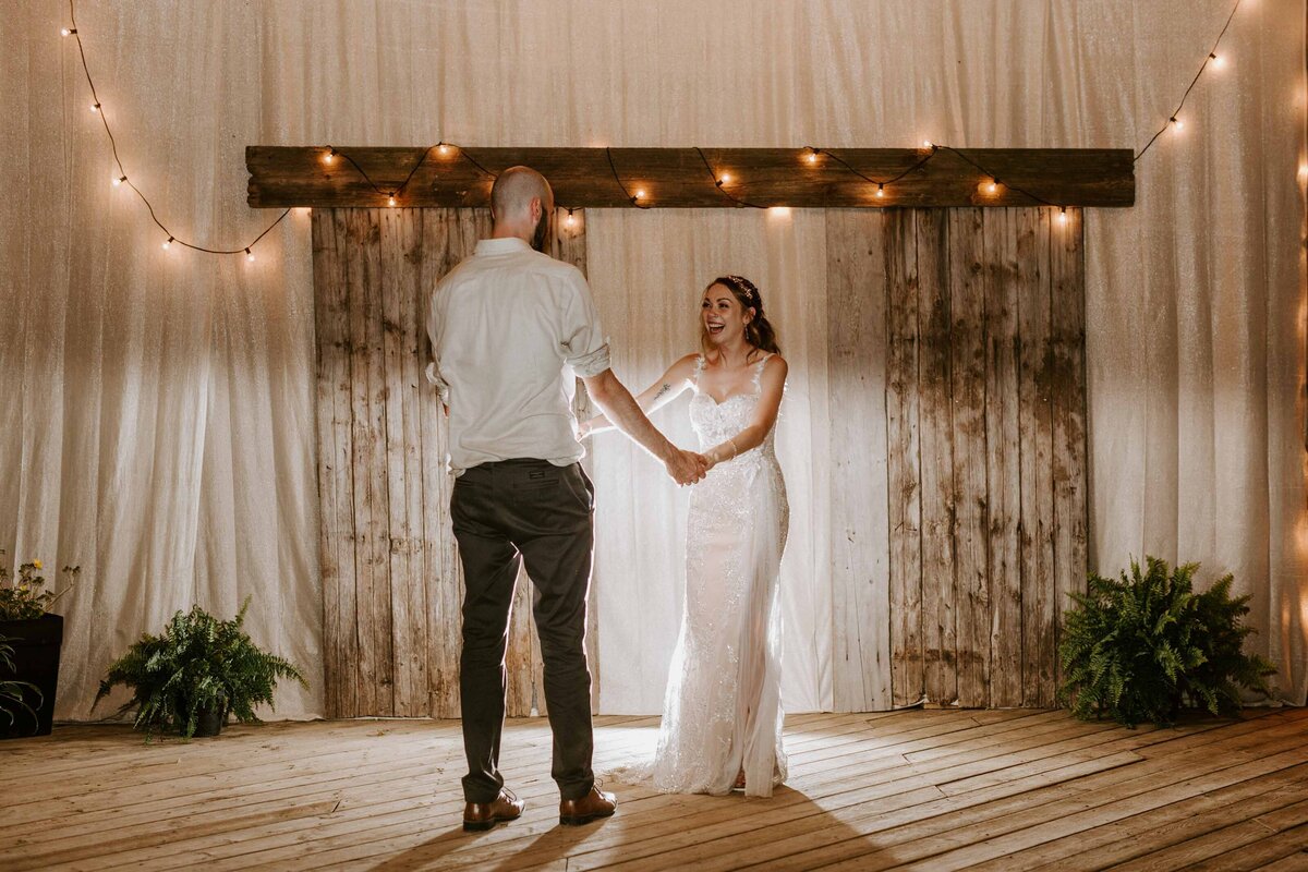 Bride and groom share their first dance at their rustic farm wedding. They are holding hands and smiling with a rustic wooden arbour behind them.