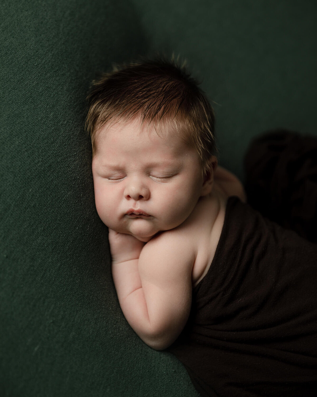 Newborn boy with lots of light brown hair laying on his stomach with a dark brown wrap and green background.
