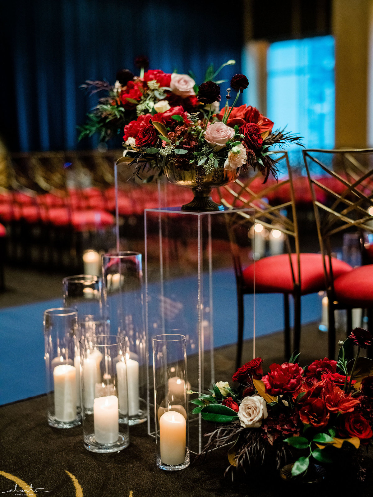 Aisle entrance decor with lucite flower stand, red winter flowers, hurricane candles, and Chameleon chairs with red seat cushion