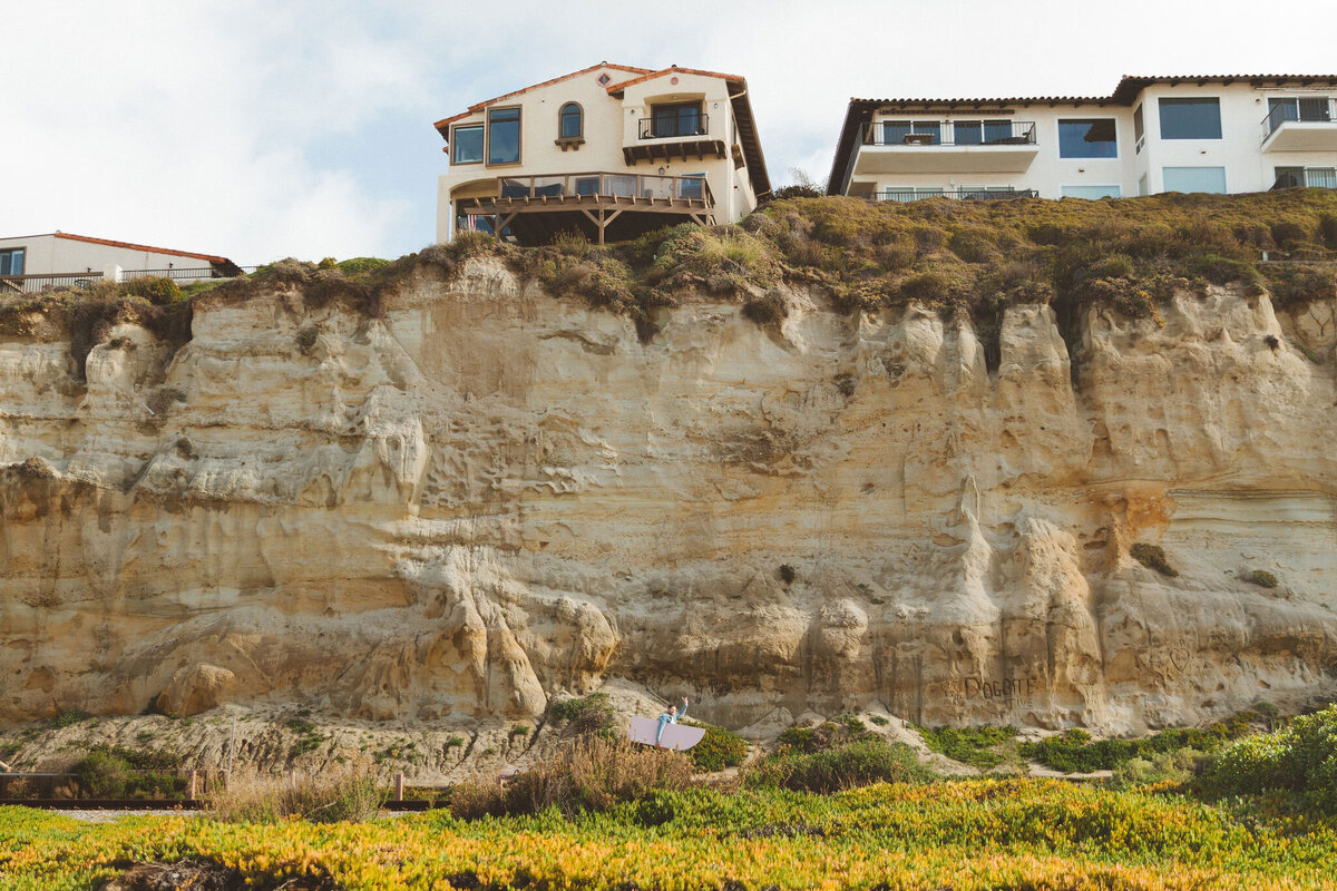 Design engineer Jake Jones setting up custom backdrops by Shindig Social against a rock formation in San Clemente, California. The scene highlights Shindig Social's service expansion to Orange County, San Diego, Los Angeles, and Palm Springs. The text reads 'now serving' with the listed regions, showcasing the company's commitment to providing bespoke event backdrops across Southern California.