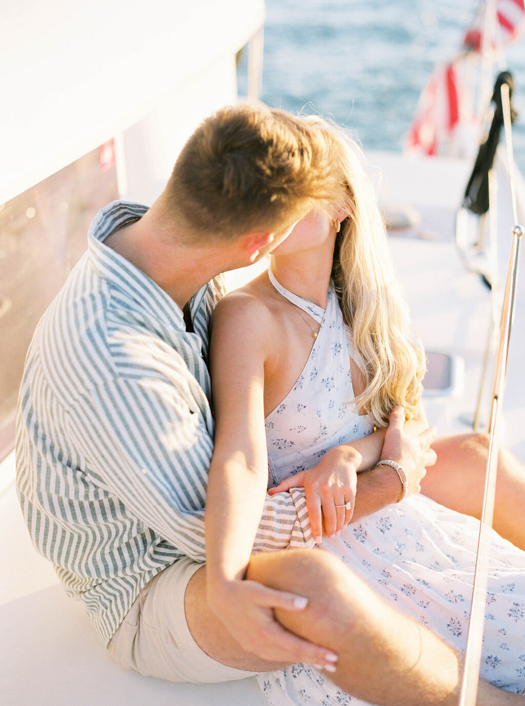 Wedding Photographer RI | A couple sits close on a boat, sharing a kiss. The woman wears a white floral dress; the man wears a striped shirt. 