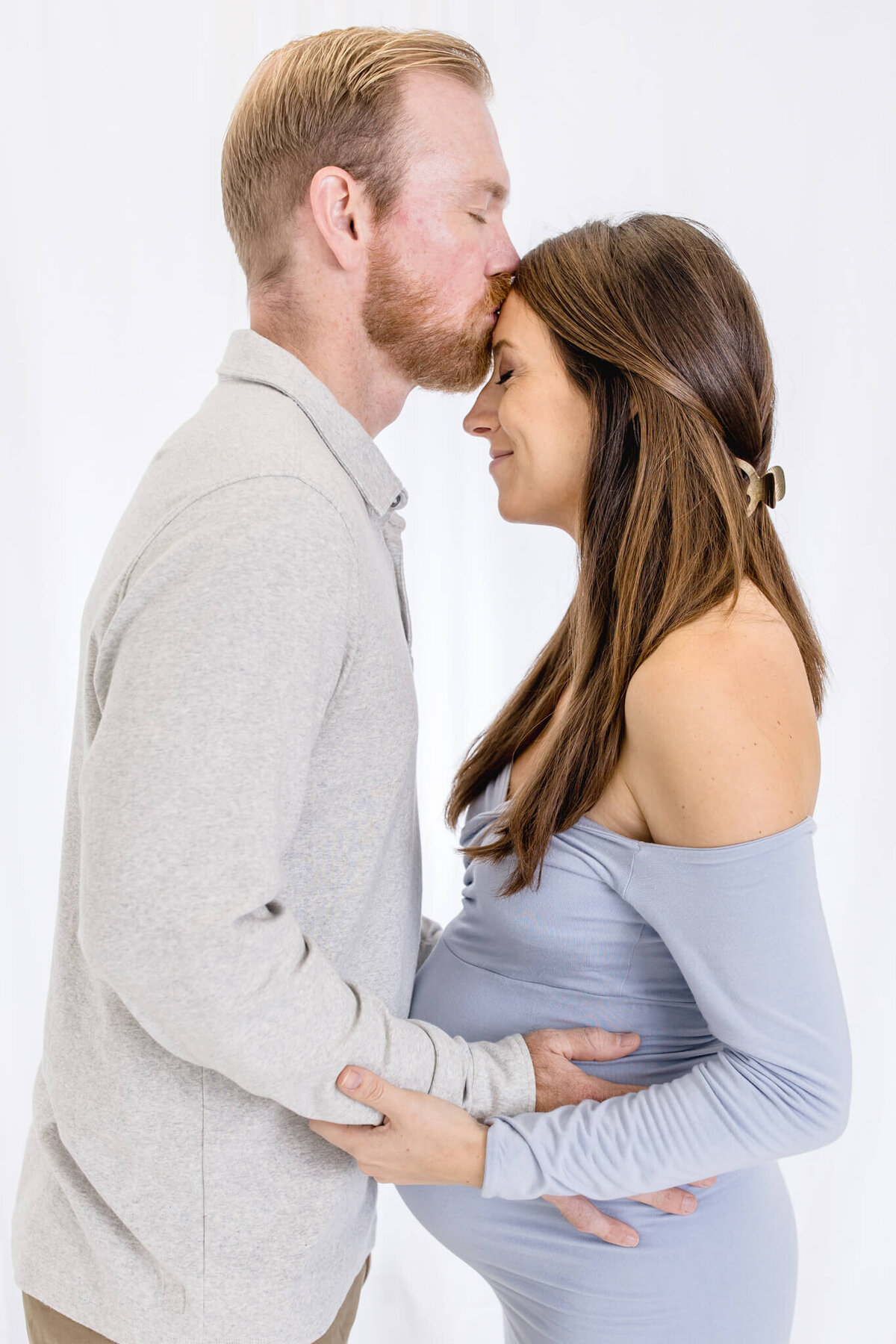 A man gently kisses the forehead of a pregnant woman, both standing in profile against a white background. The woman cradles her belly with her hands, wearing a blue off-shoulder dress. The man wears a light gray sweater.
