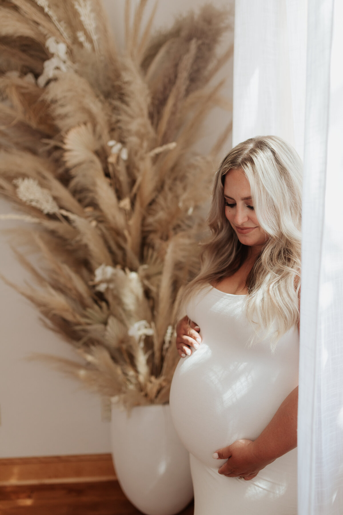 Pregnant mother standing against a window with white curtains, wearing a white dress holding baby bump