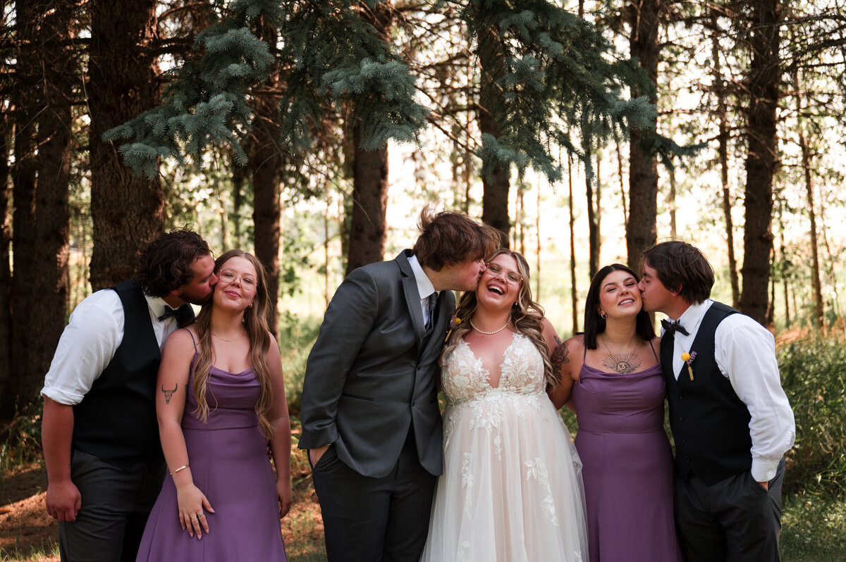 Wedding party with 3 couples all men are kissing the women on the cheek, smiling and laughing in wedding photo