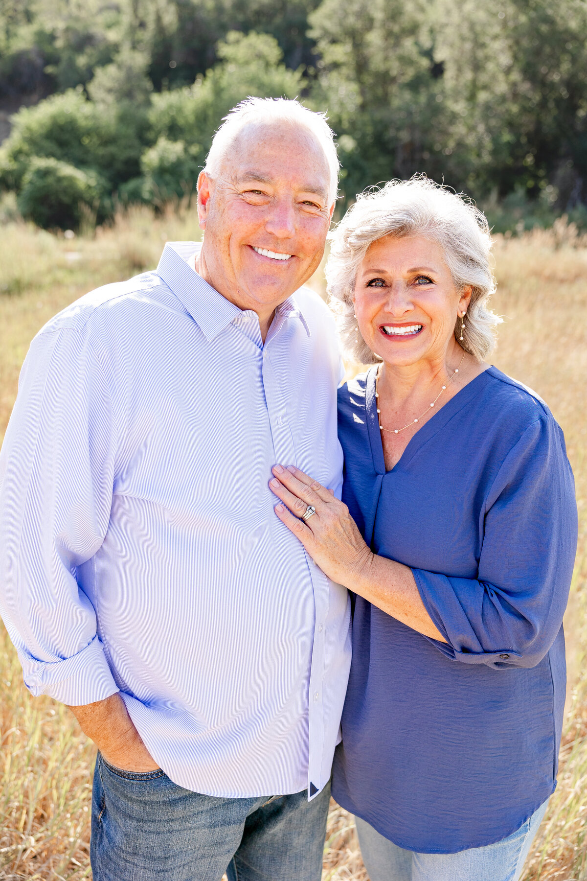 An elderly couple smiling warmly at the camera, dressed in matching blue outfits, standing in a golden field.