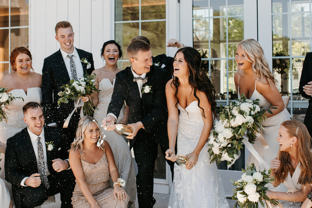 Bride and groom popping champagne while their bridal party celebrates around them