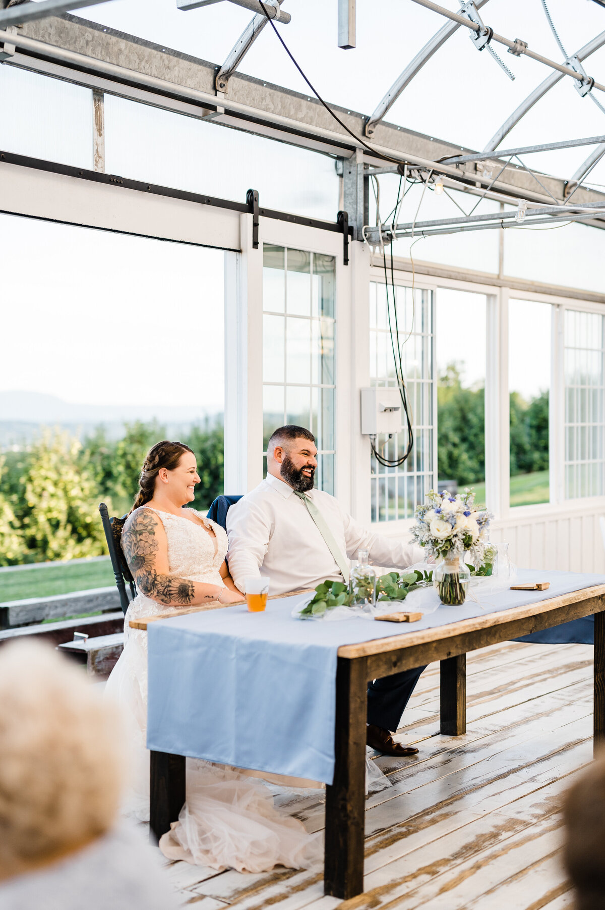 wedding reception at showalter's orchard with bride and groom sitting at a reception table at greenhouse reception hall as they laugh with their wedding guests at toast speeches
