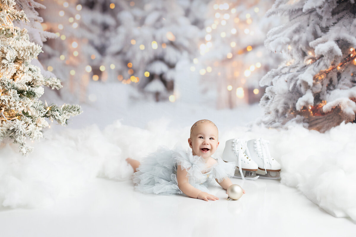 Smiling baby in a soft blue tulle dress holding a gold ornament, lying between snow-dusted Christmas trees with ice skates nearby, in a magical winter scene created by a Dallas milestone photographer.