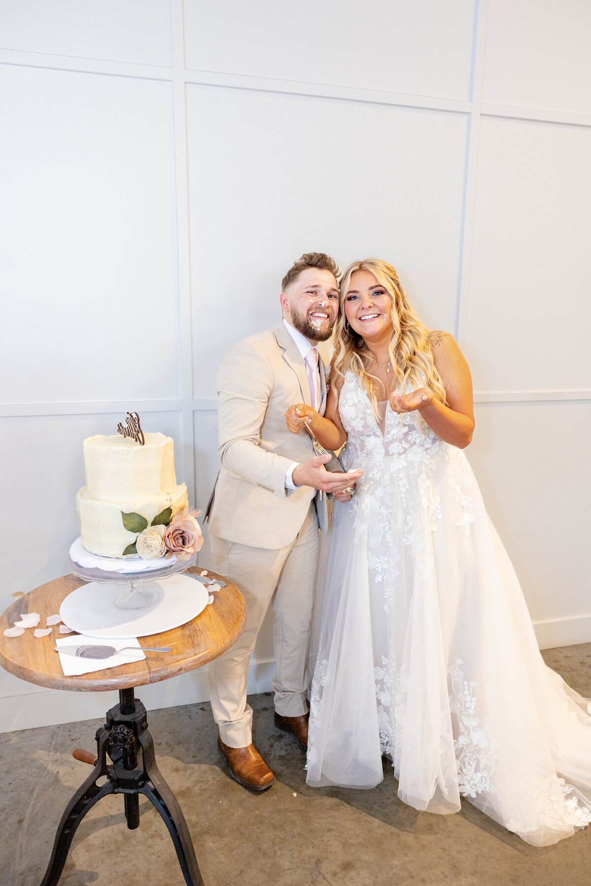 The bride and groom stand next to their wedding cake, laughing and showing traces of cake on their hands and faces.