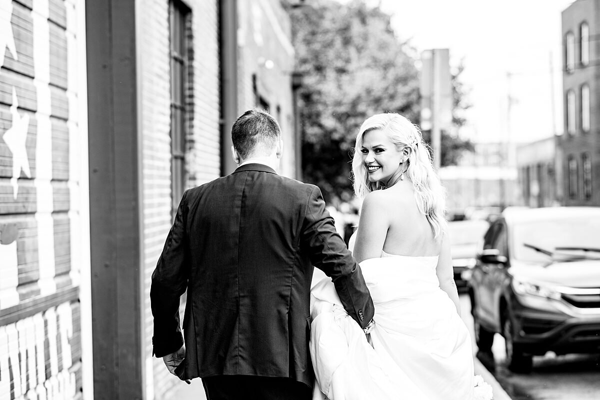 Black and white photo of the bride and groom walking away from the camera down a city street. The groom is wearing a dark suit and the bride looks over her shoulder at the camera smiling. She is wearing a strapless dress with a long flowing train that the groom is helping her carry.