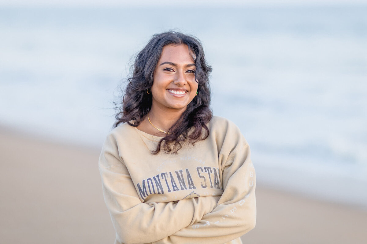 A person with long, curly hair smiles while standing on a beach. They are wearing a beige sweatshirt with Montana State written on it. The ocean is blurred in the background.