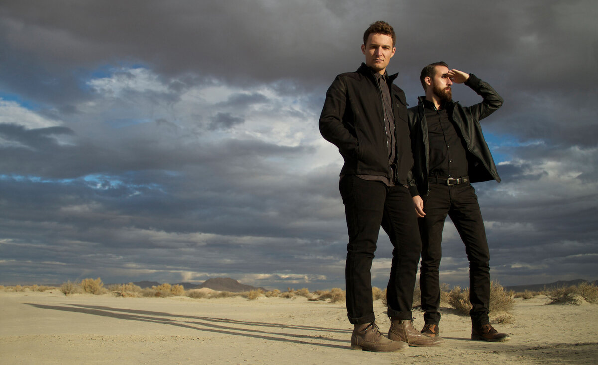 Musical duo portrait Polarcode standing in desert  one member hand up shielding light from eyes stormy clouds behind