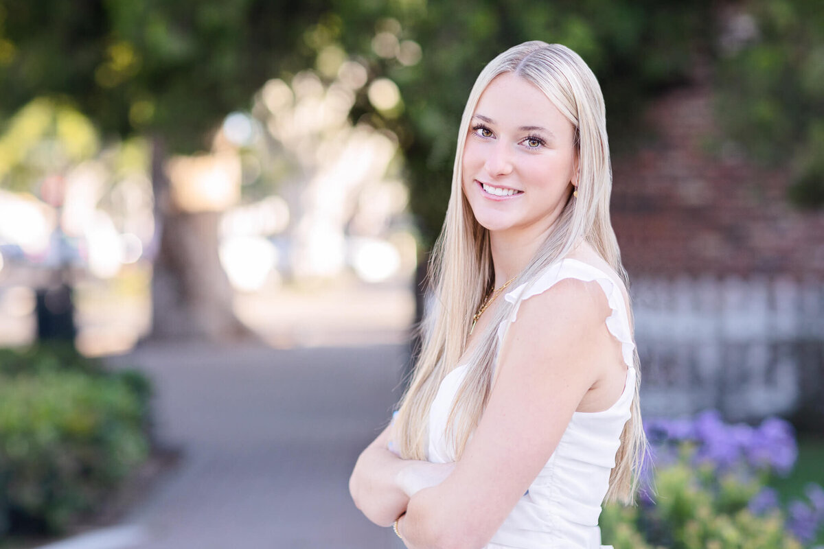 A young woman with long blonde hair stands outdoors, smiling with her arms crossed. She is wearing a white sleeveless top, and the background is softly blurred, featuring greenery and a pathway.