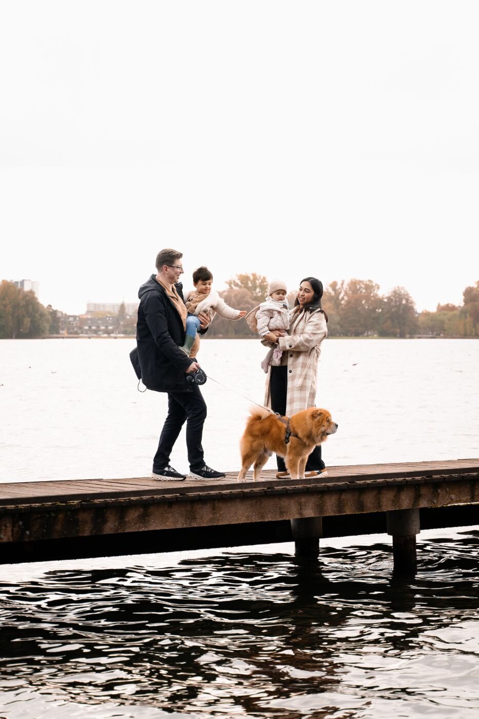 Family of four with dog standing on a wooden pier by the lake on a cloudy autumn day.